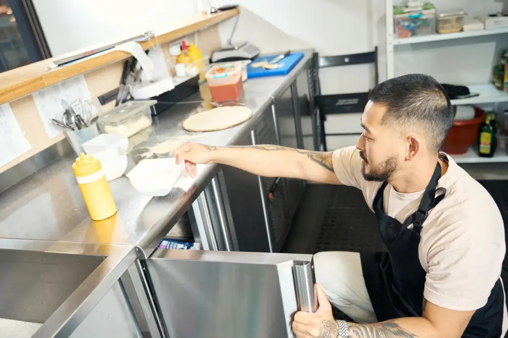 Professional cook in apron reaching into commercial undercounter refrigerator
